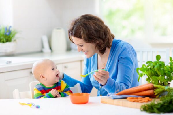Fröhliche Mutter füttert lachendes Baby mit biologischer Karottenbabynahrung in einer hellen Küche
