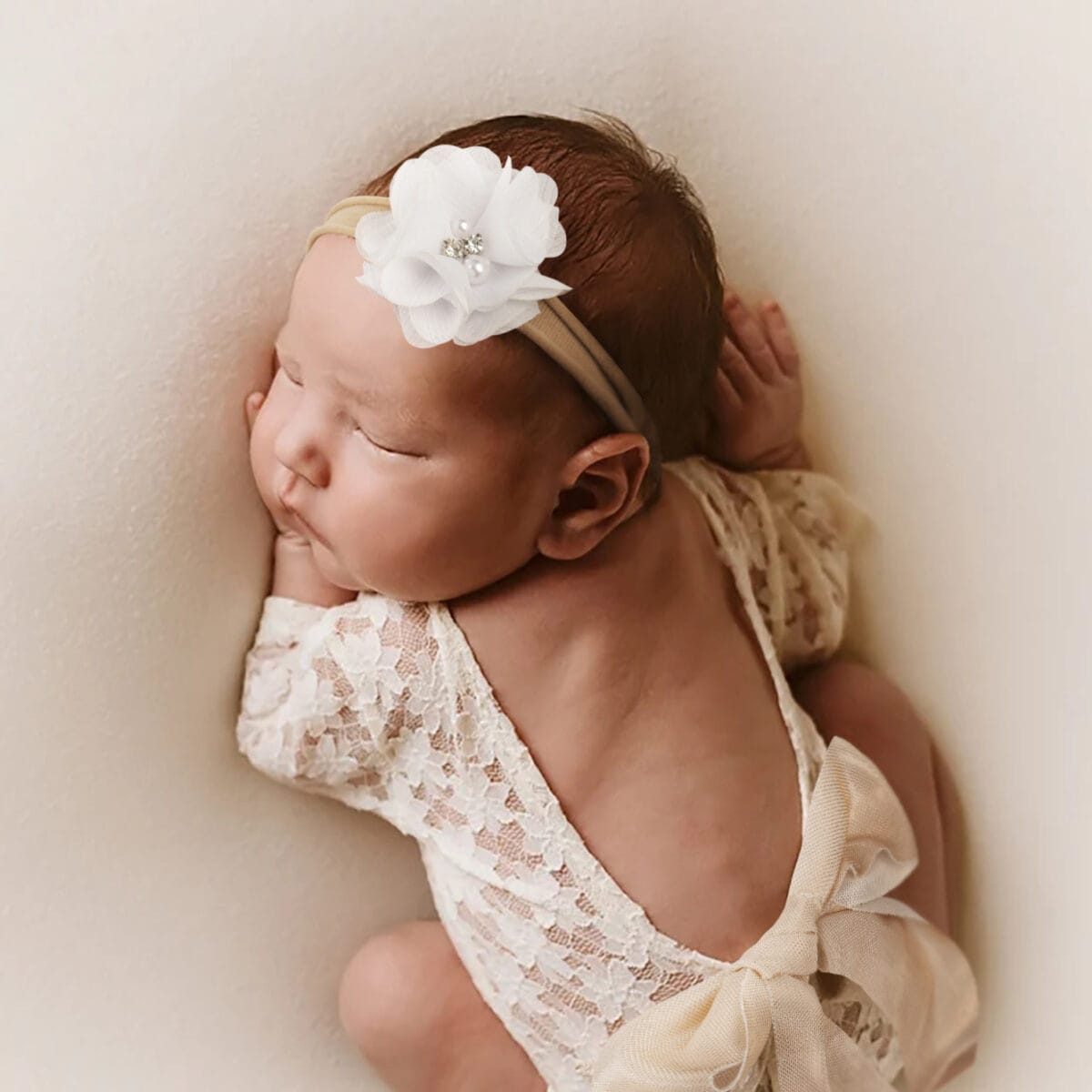 Neugeborenes schlummert friedlich im Spitzen-Strampler mit rückenfreiem Design und weißem Blumen-Haarband, perfekt für Neugeborenenfotografie.