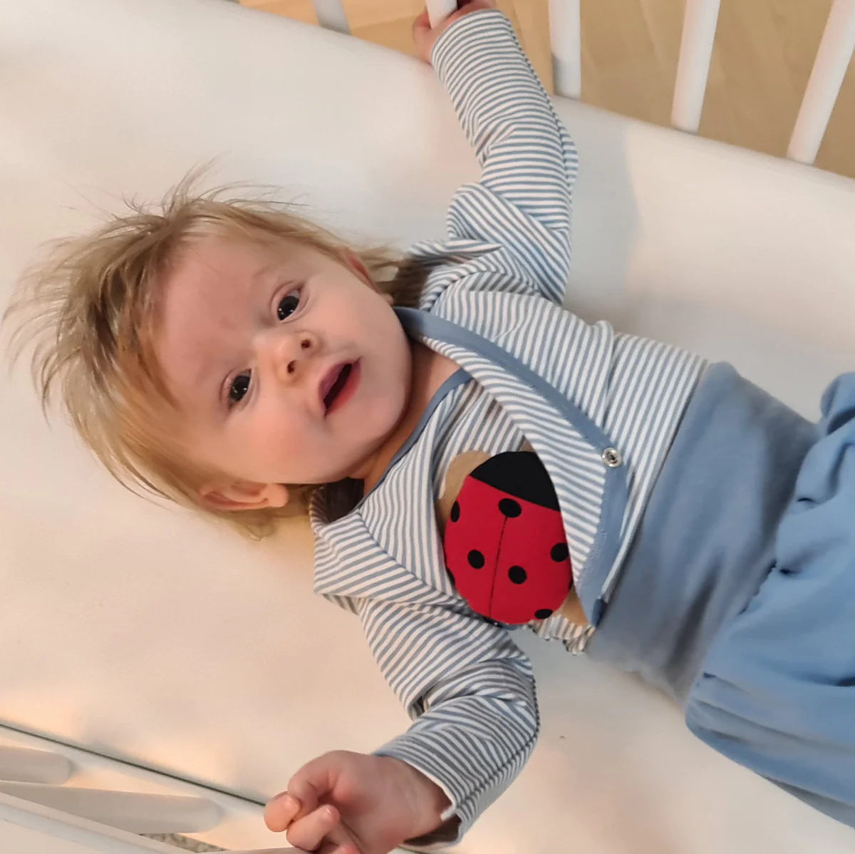 Lächelndes Baby im blau-gestreiften Body mit Marienkäfer-Applikation liegt entspannt im Kinderbett.